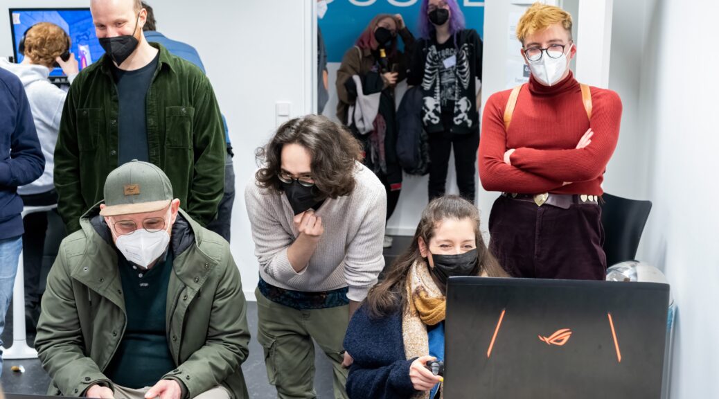 A photo taken by Julian Dasgupta at the & Play event in April 2023 showing two people smiling while playing a game in front of laptops with some amused observers standing behind them. A third person (presumably one of the developers of the game being played) is talking with one of the players. Everyone's wearing a mask.